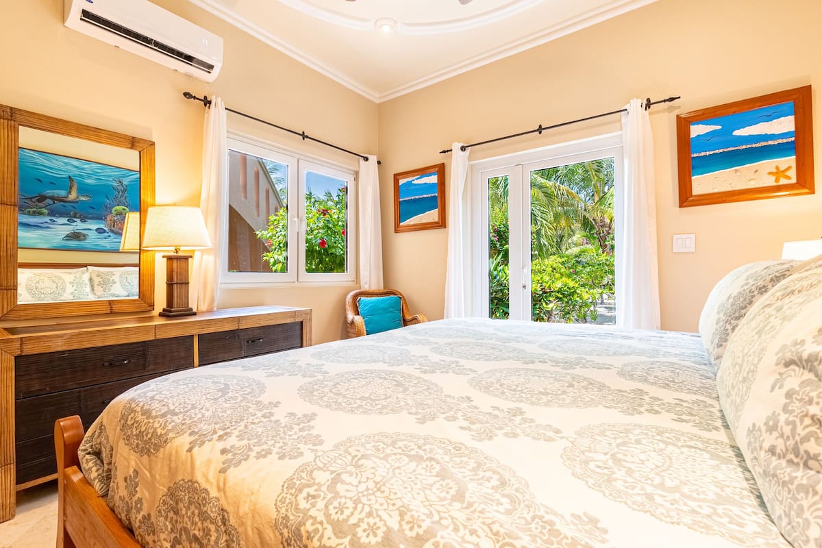 A bedroom with a large bed, a wooden dresser, a desk with a lamp, and a window with curtains.