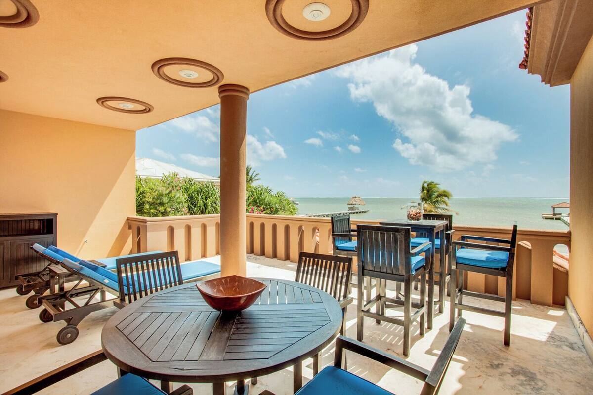 A sunlit outdoor seating area with a view of the ocean, featuring a round table, chairs, and a wooden bench.