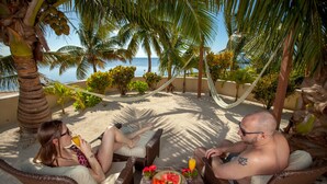 Escena en la playa con mobiliario de mimbre, una hamaca y una mesa con frutas y bebidas.