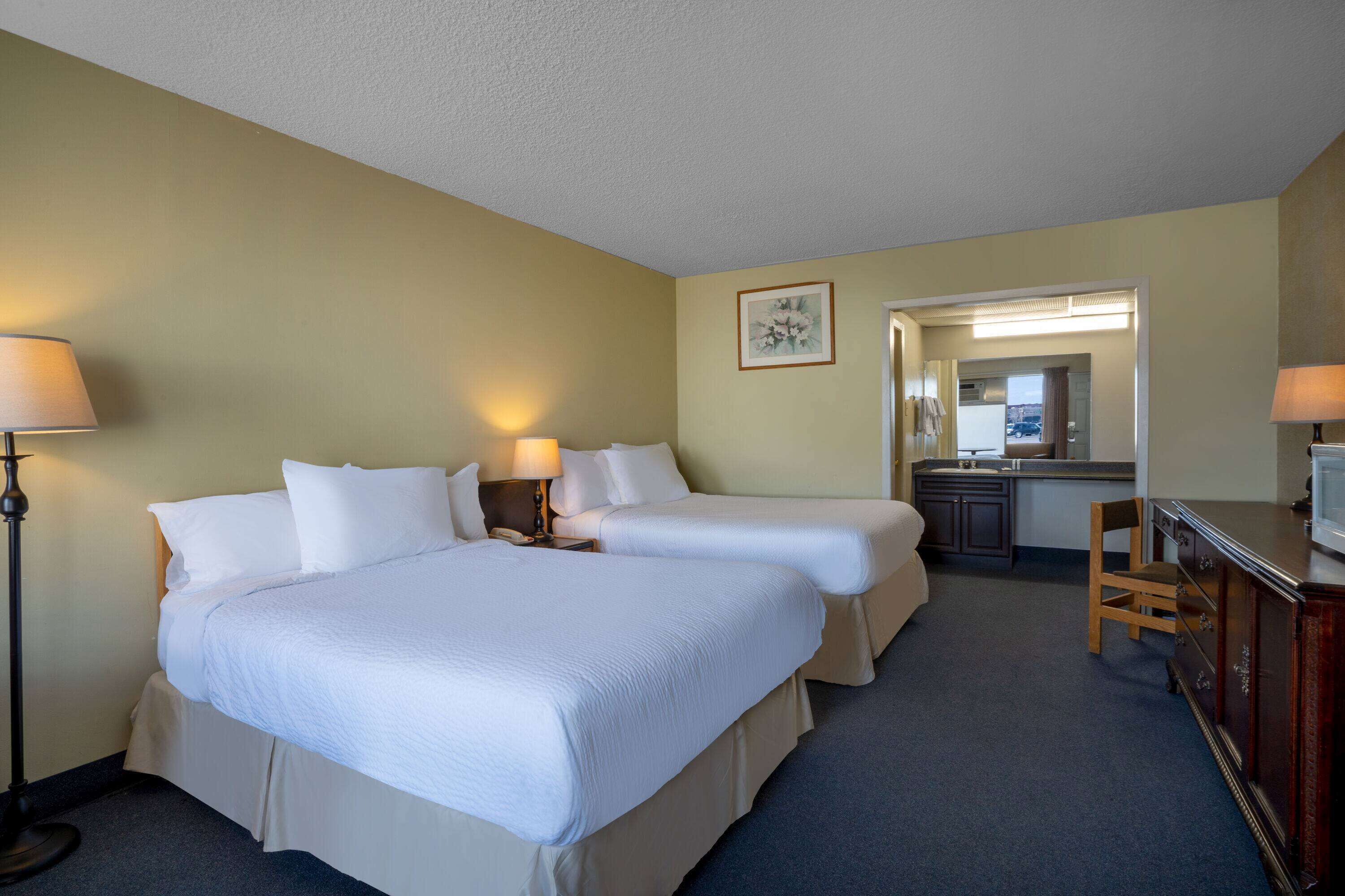 A hotel room with two beds, a desk, a chair, and a kitchenette.