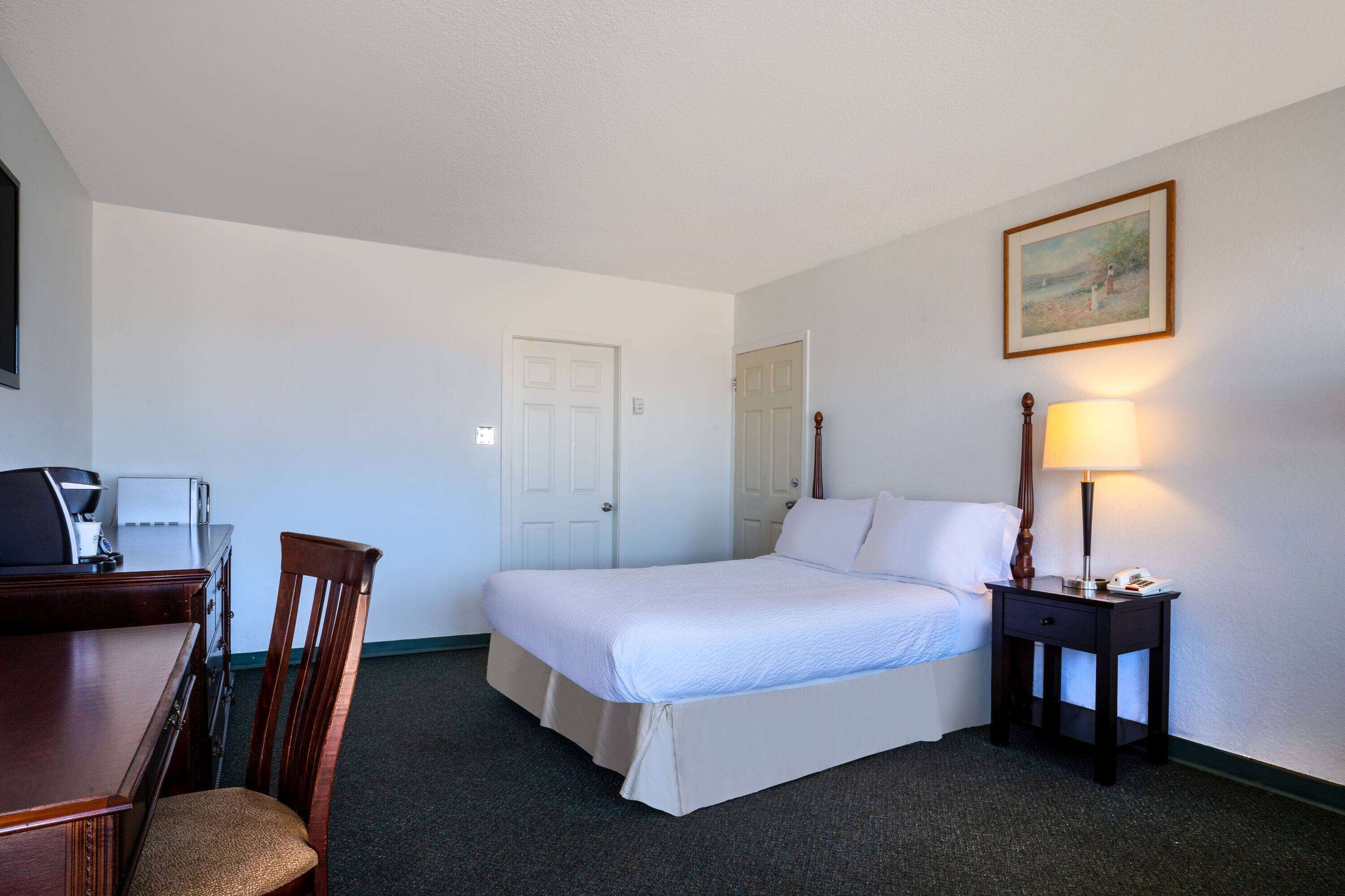 Habitación de hotel con cama, escritorio, silla, lámpara y un cuadro en la pared.