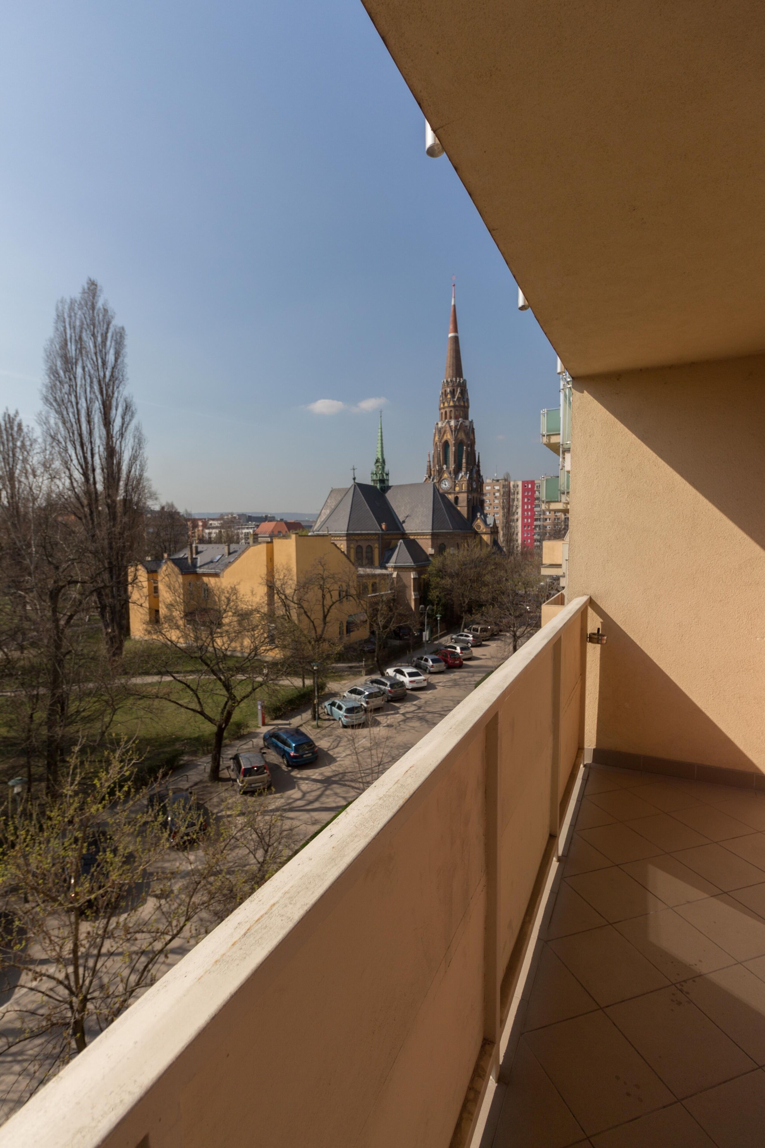 A balcony overlooking a parking lot with several cars, a church with a tall spire, and a clear blue sky.