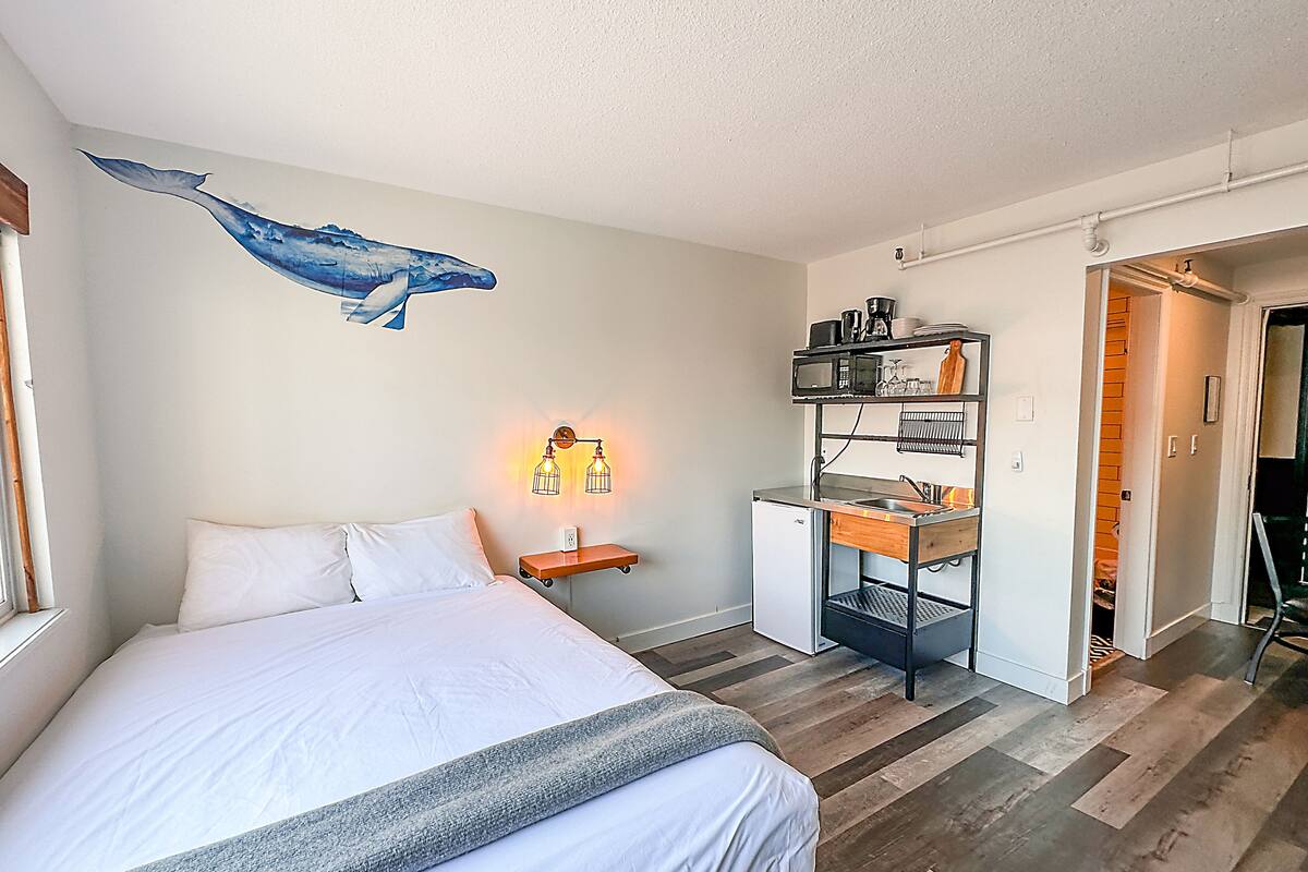 A bedroom with a bed, a wall-mounted shelf with kitchen items, and a door leading to another room.
