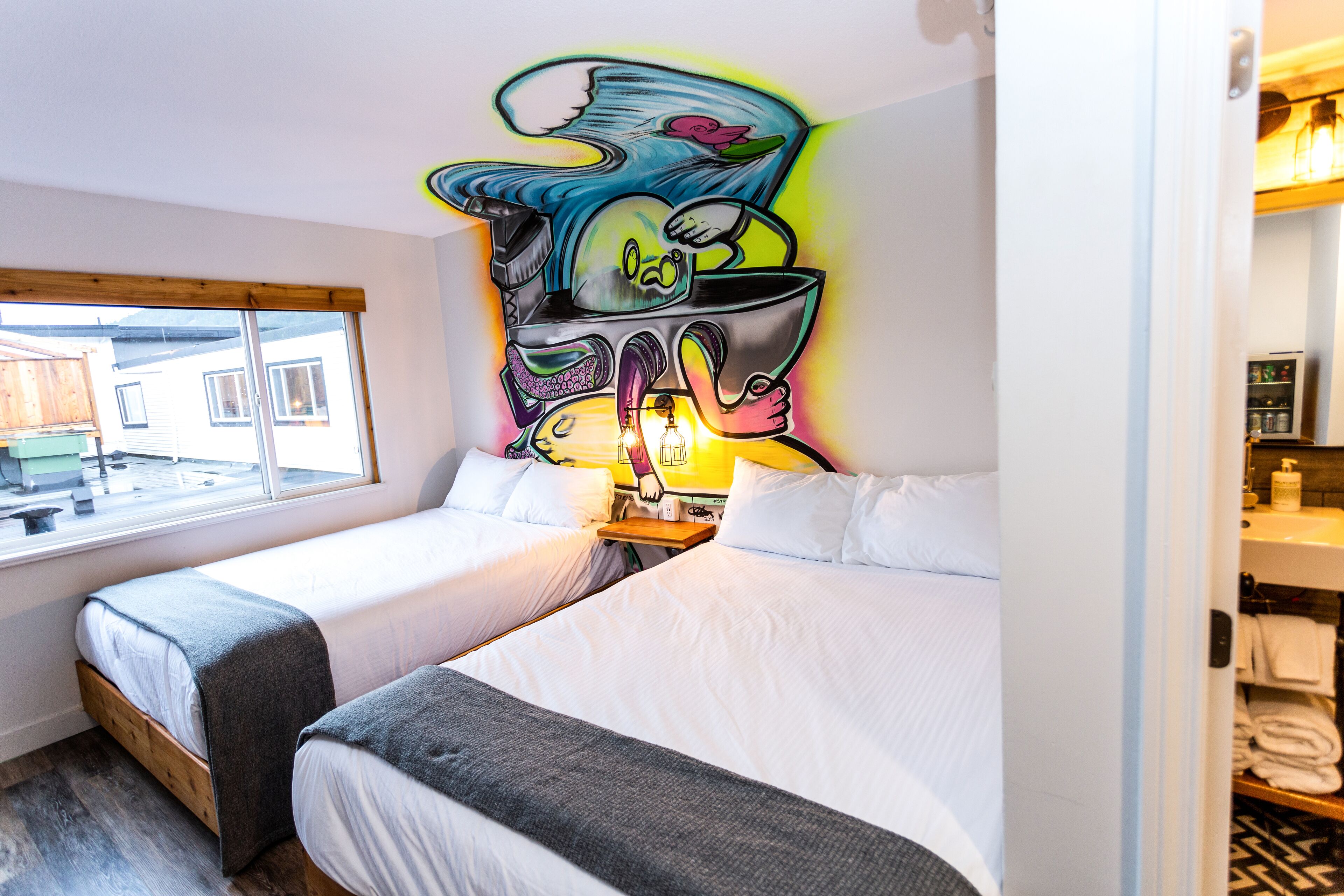 A hotel room with two beds, a large window, and a colorful mural on the wall.