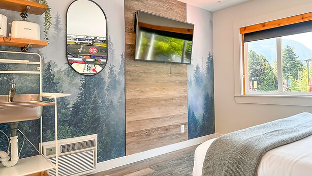 A modern bedroom with a bed, a window with a view of trees, a wall-mounted TV, and a mirror. - Crash Hotel Squamish (Squamish)