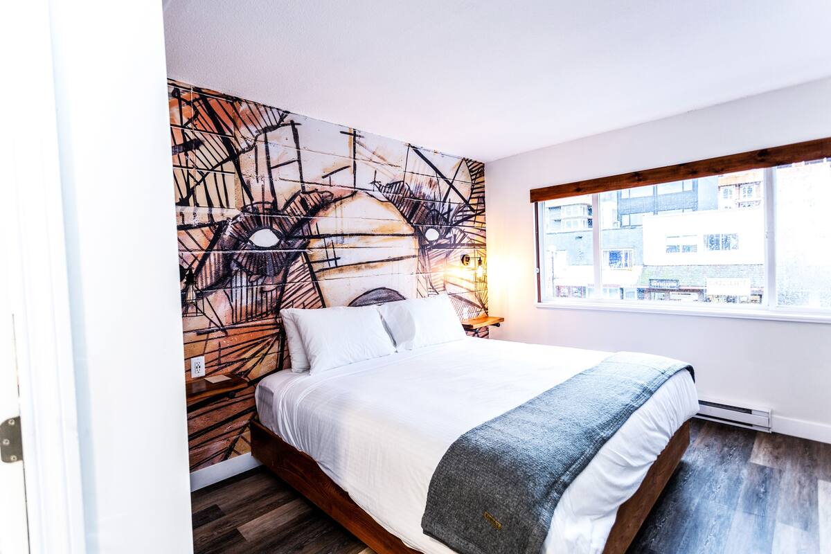 A bedroom with a large bed, a wall mural, a window with a view of buildings, and a wooden floor.
