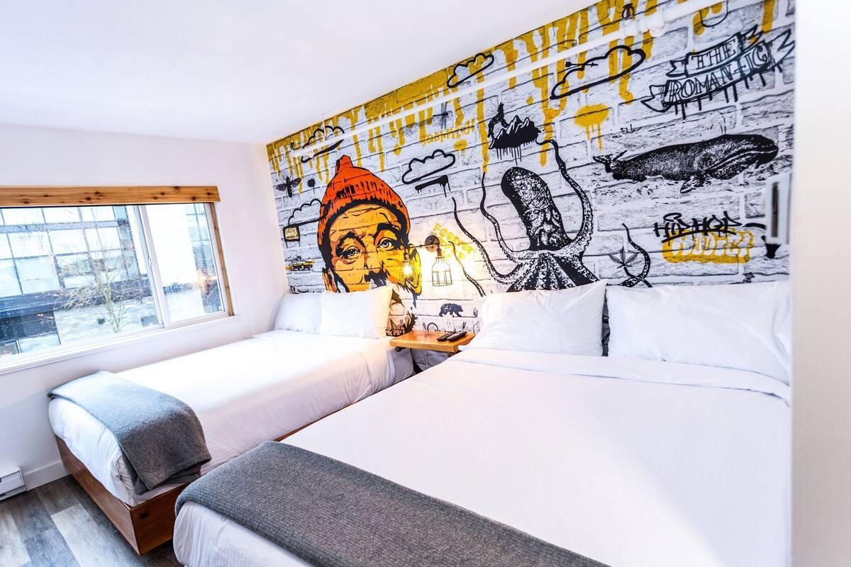 A hotel room with two beds, a large mural on the wall, and a window with a view of buildings.