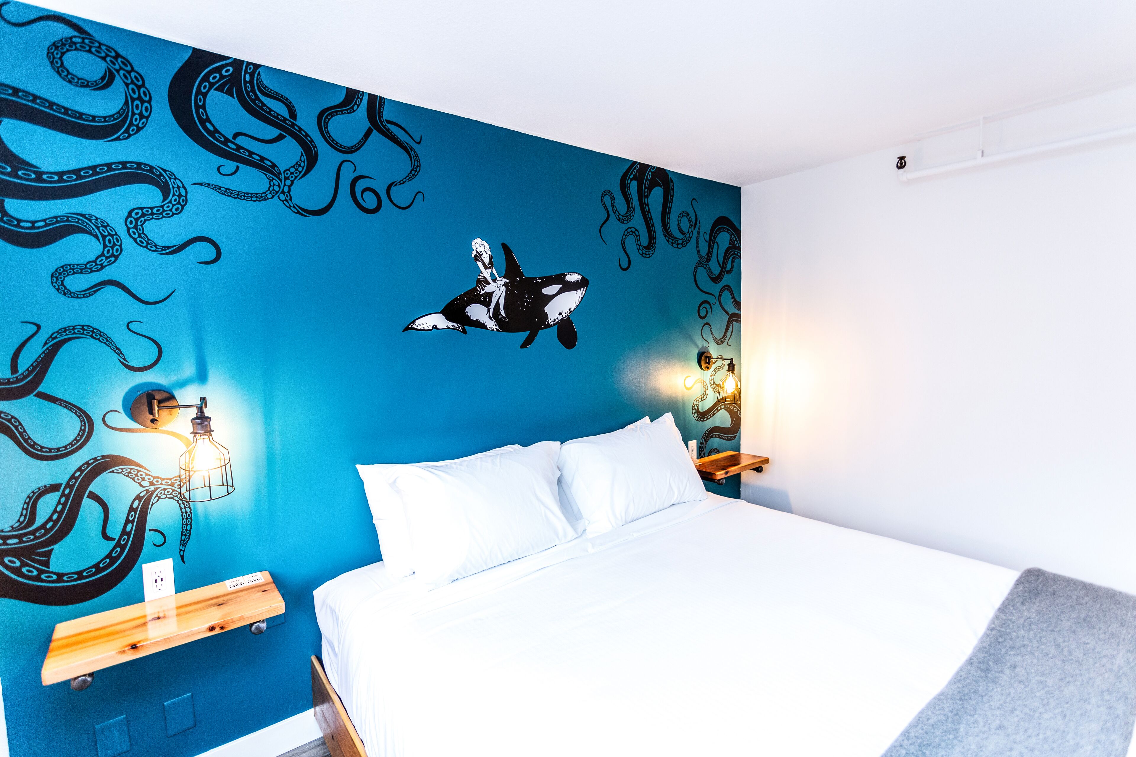 Un dormitorio con pared azul decorada con un mural de pulpo y ballena, una cama con ropa de cama blanca y una mesita de noche con una lámpara.