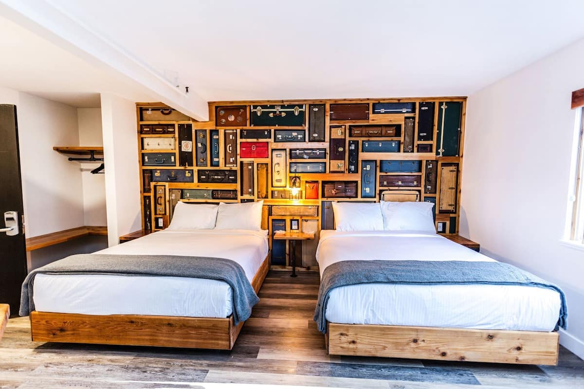 A room with two wooden beds, a wall of suitcases, and a small table with a lamp.