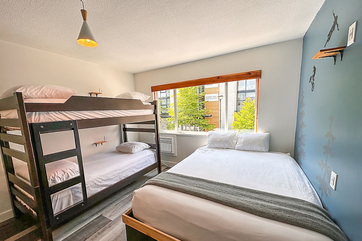 A room with bunk beds, a single bed, a window with a view of trees, and a hanging light fixture.