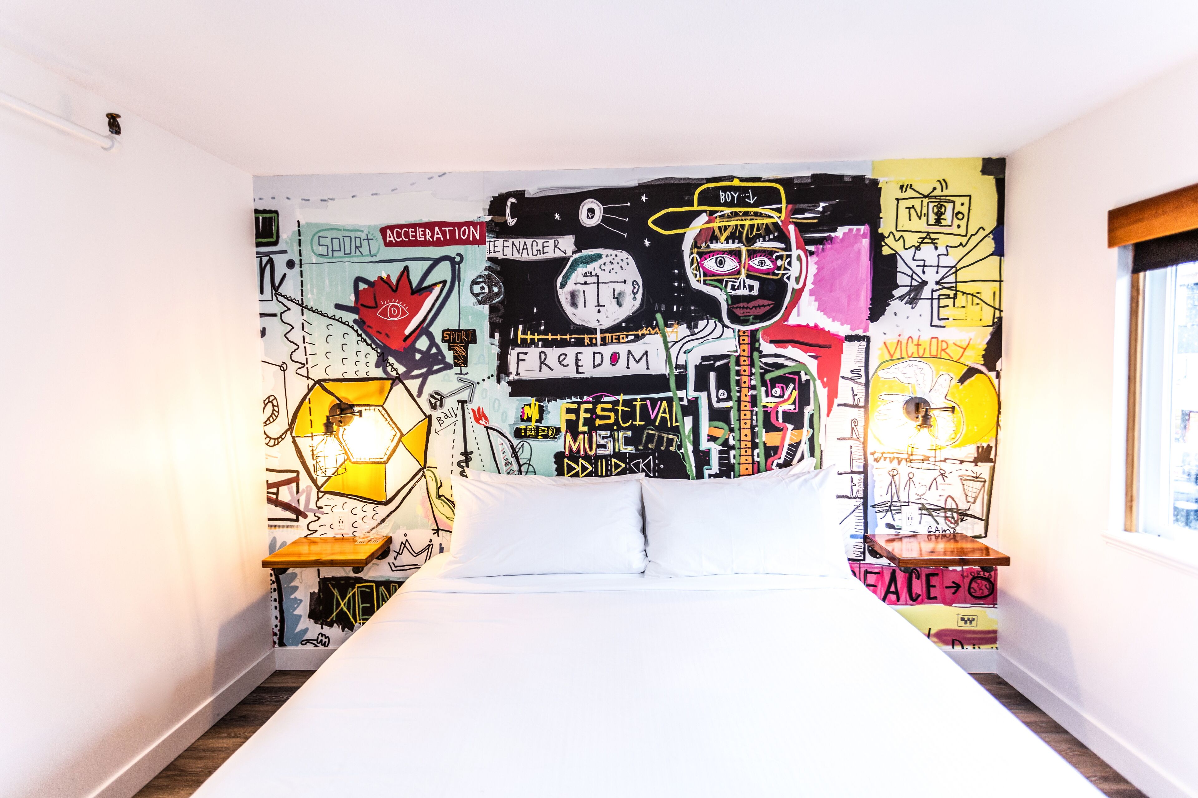 A bedroom with a bed centered in the room, a wall mural with various words and symbols, and a window on the right side.