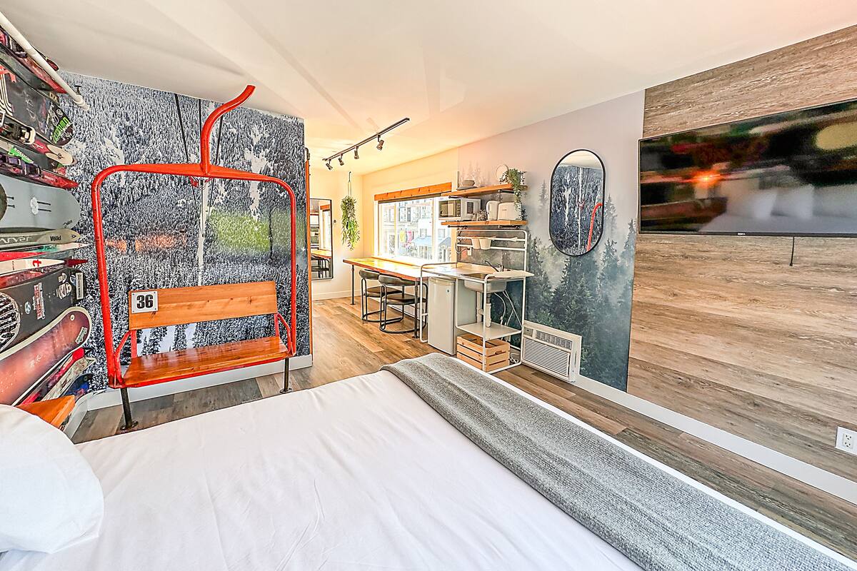 A modern hotel room with a bed, a desk with a chair, a small kitchenette, and a large window.