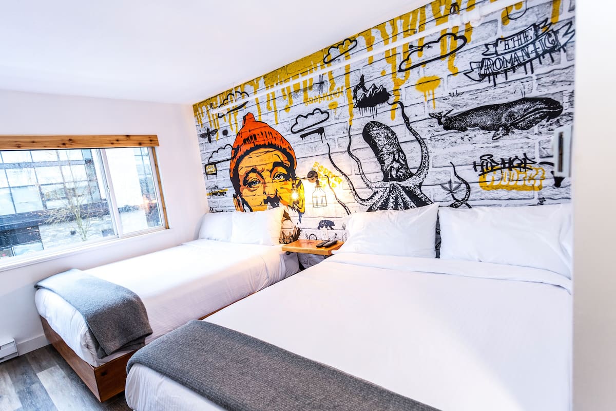 A hotel room with two beds, a large mural on the wall, and a window with a view of buildings.