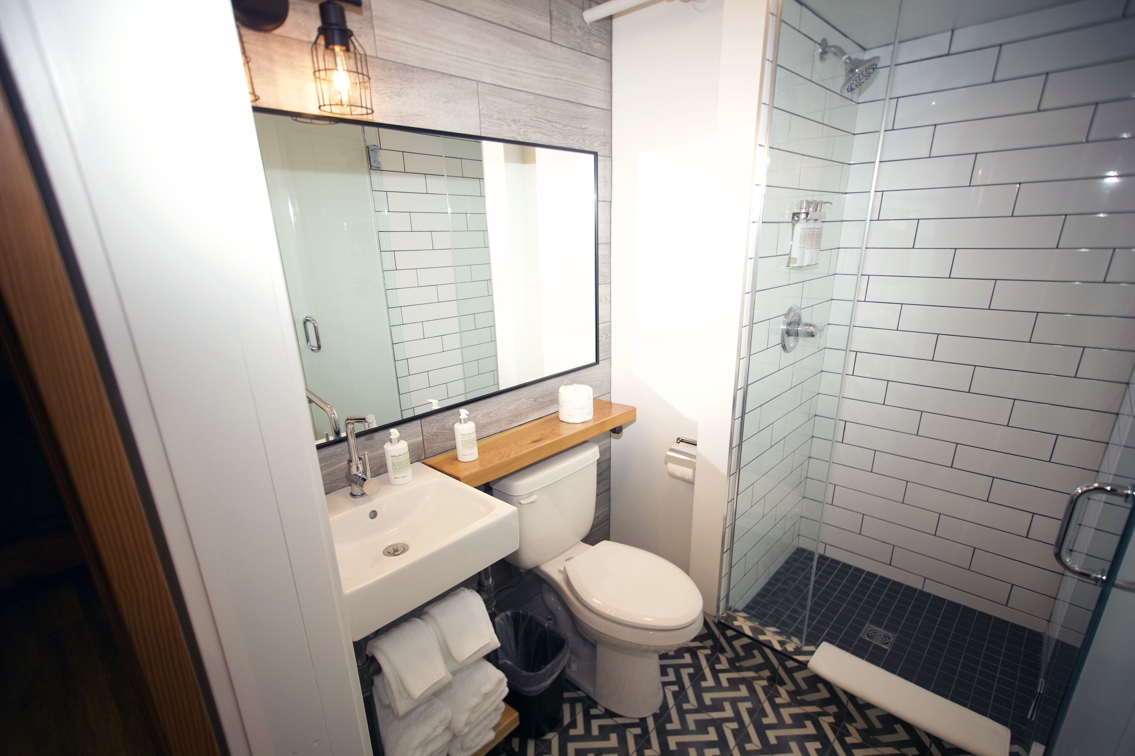 A bathroom with a white sink, toilet, and shower. There is a mirror above the sink and a wooden shelf with toiletries. The floor has a black and white patterned tile.