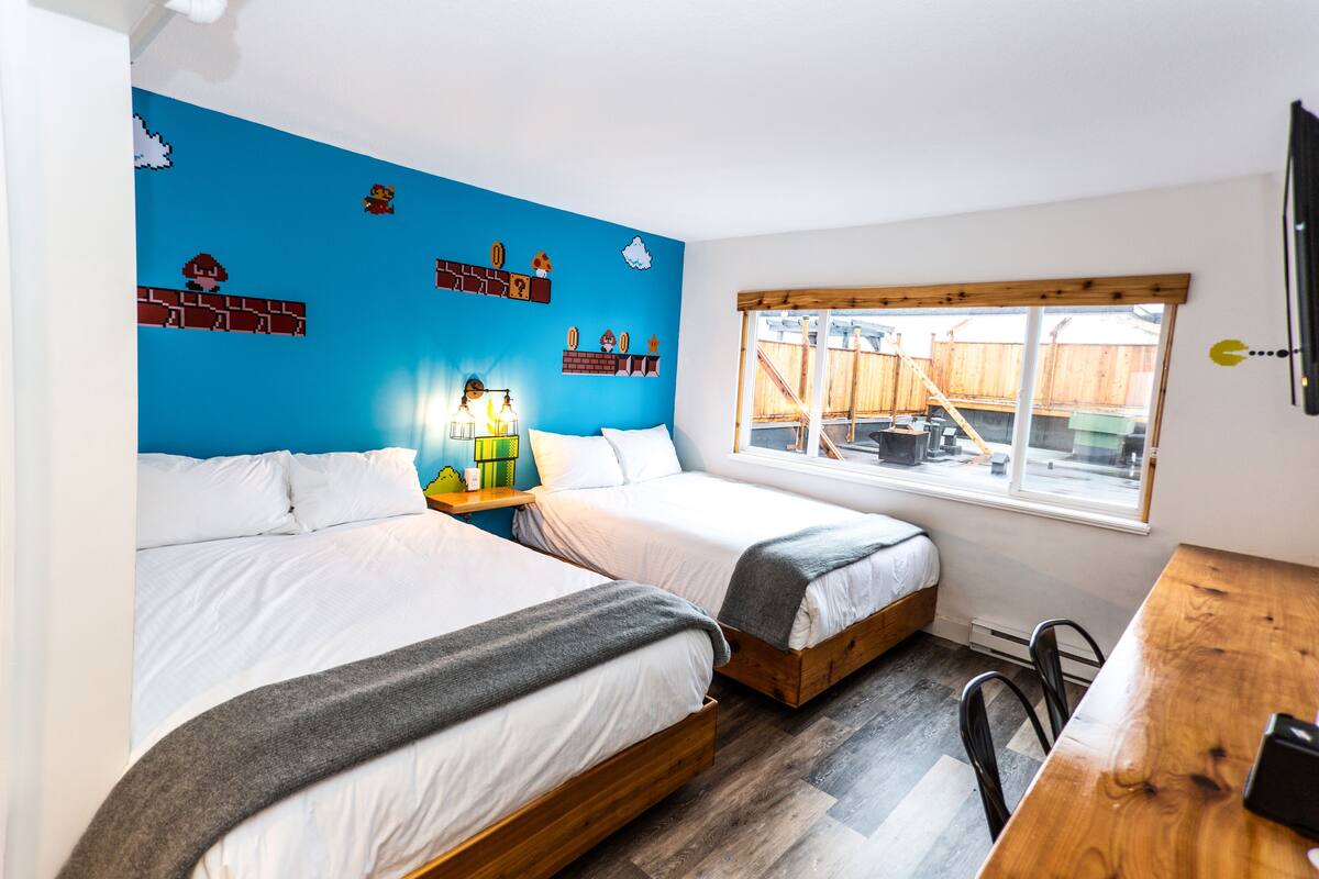 A room with two beds, a wooden table, a window with a view of a backyard, and a wall decorated with a Mario theme.