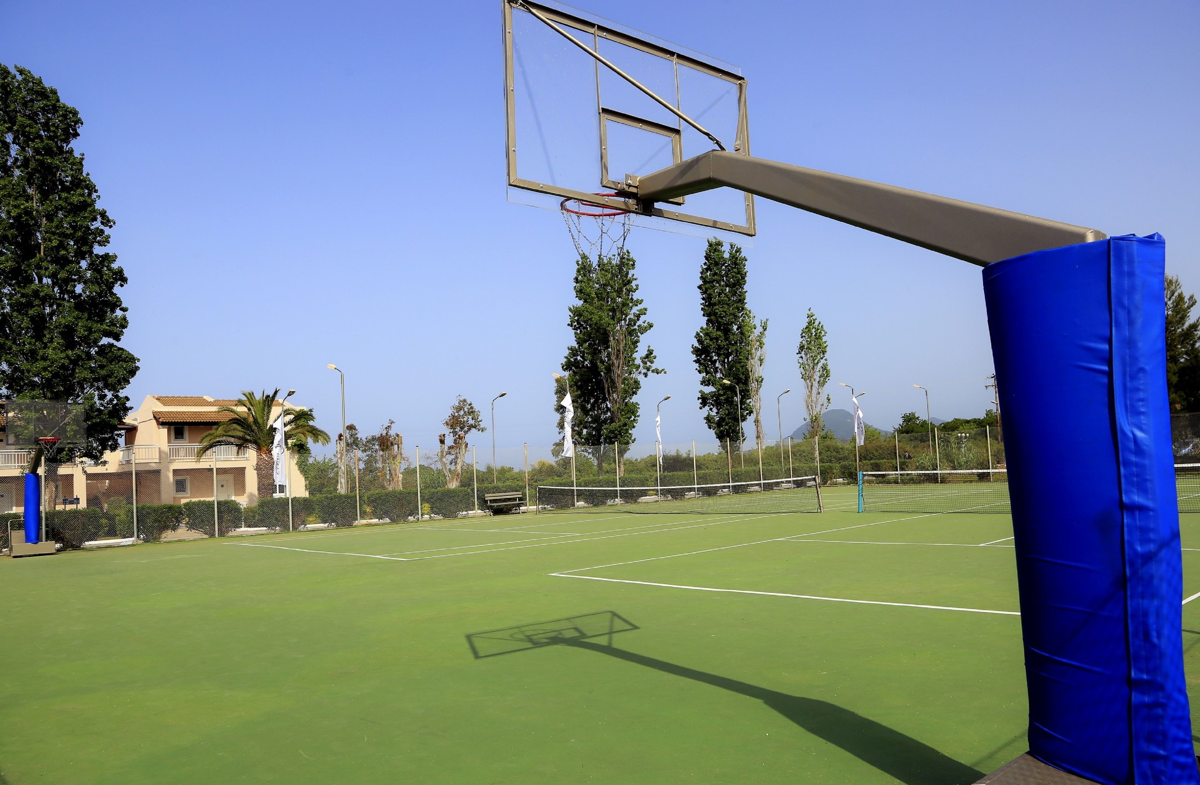 An outdoor tennis court with a net, a basketball hoop, and a blue padded area.