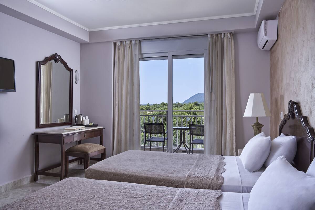 Une chambre d’hôtel équipée d’un lit, d’un bureau, d’une chaise, d’une table et d’un balcon offrant une vue sur la verdure et une montagne.
