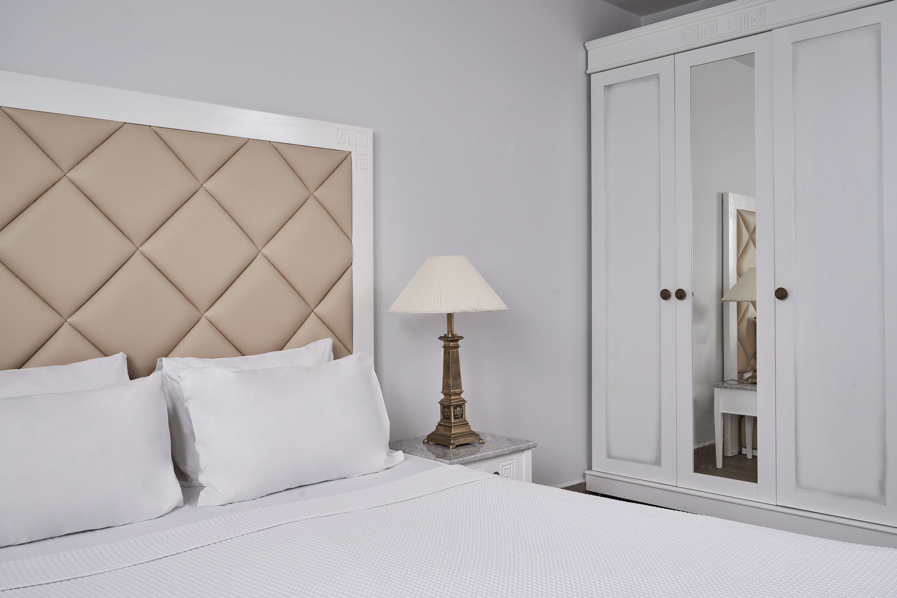 A bedroom with a tufted headboard, a bedside lamp, a wardrobe, and a mirror.