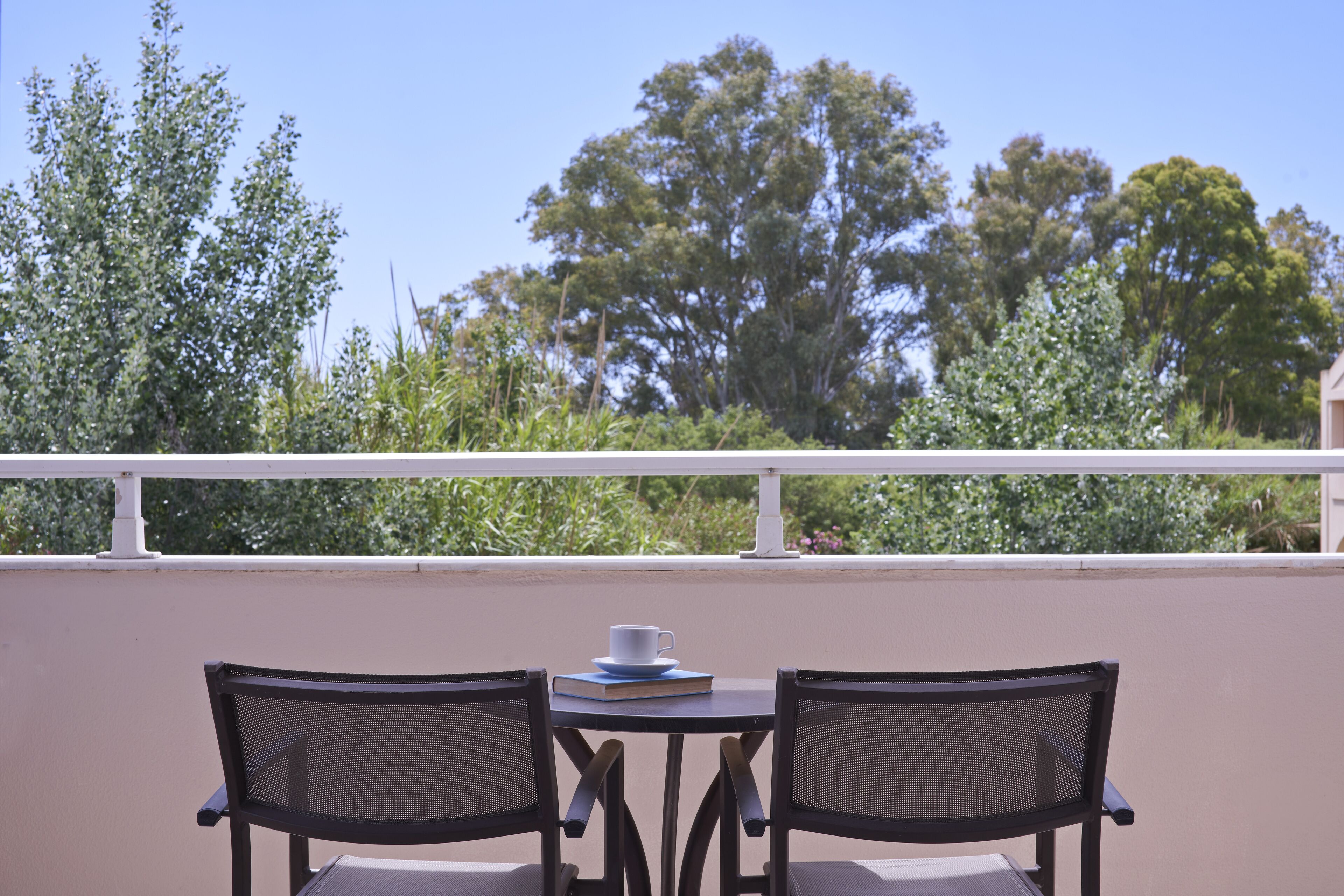 A balcony with two chairs and a table, a white railing, and a view of trees and greenery.