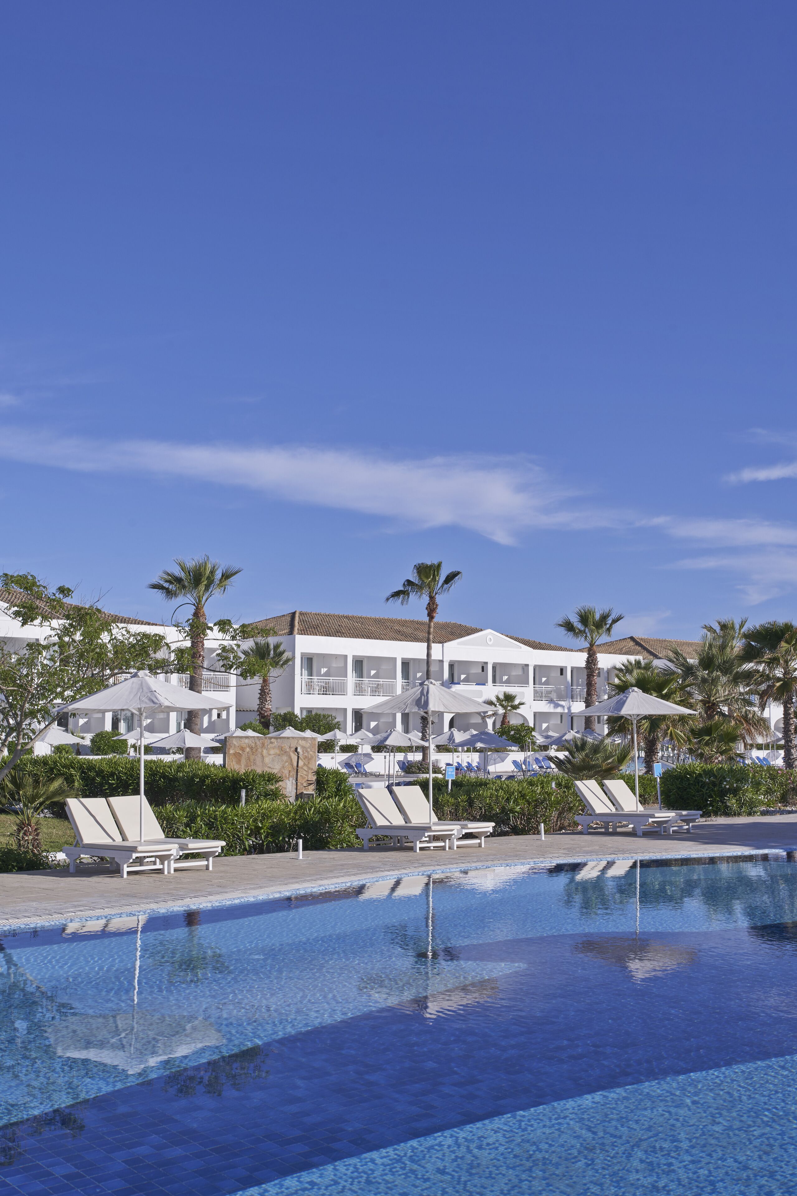 A swimming pool with lounge chairs and umbrellas, surrounded by palm trees and a building with balconies.