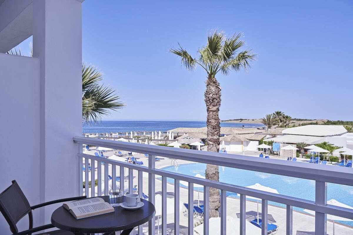 Un balcon avec vue sur une plage et une piscine, comprenant une table avec un livre et une tasse.