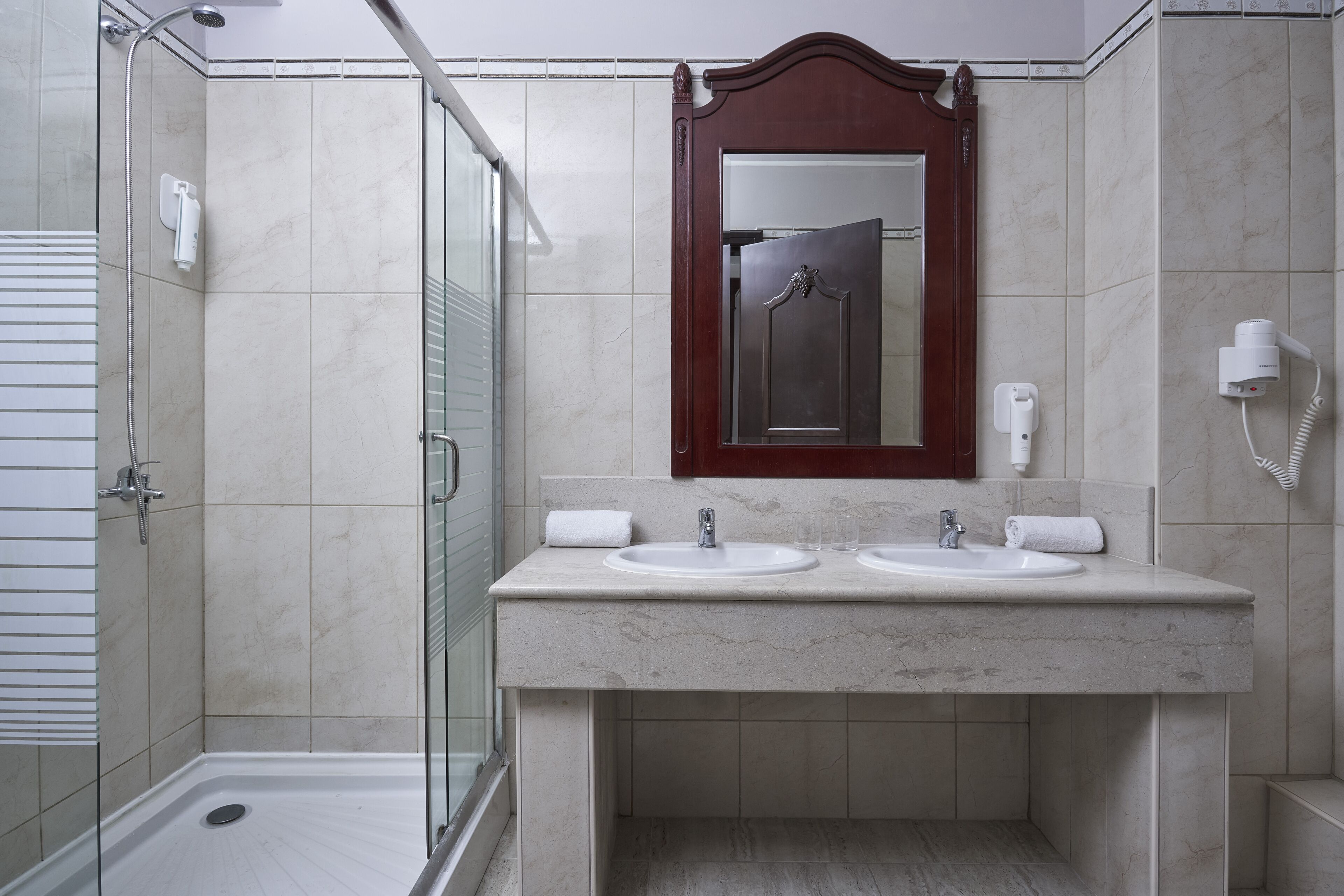 A bathroom with a glass-enclosed shower, two sinks, a mirror, and a hairdryer on the wall.