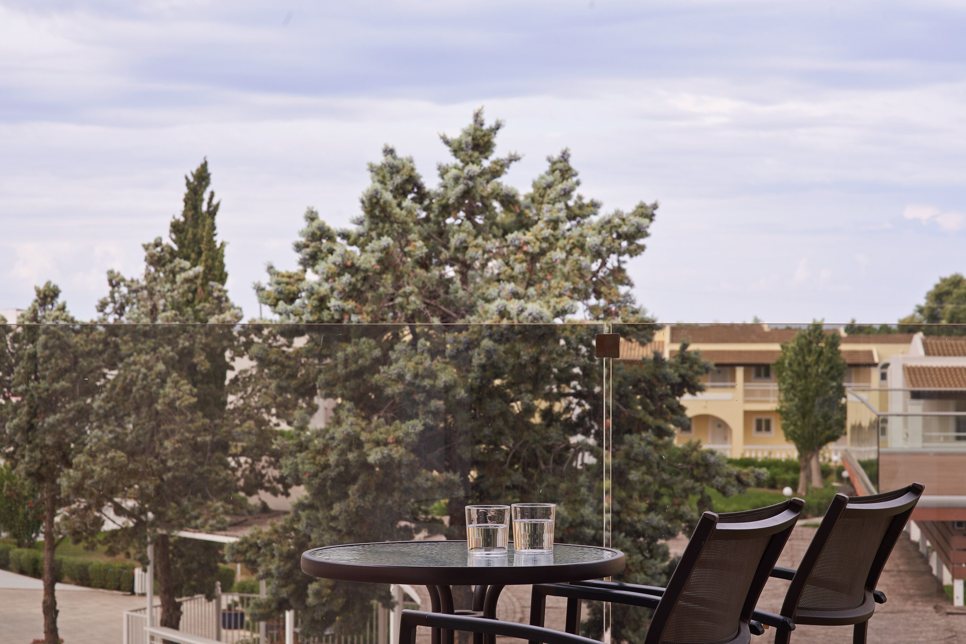 A round glass table with three clear glasses, two dark brown chairs, and a view of trees and buildings in the background.