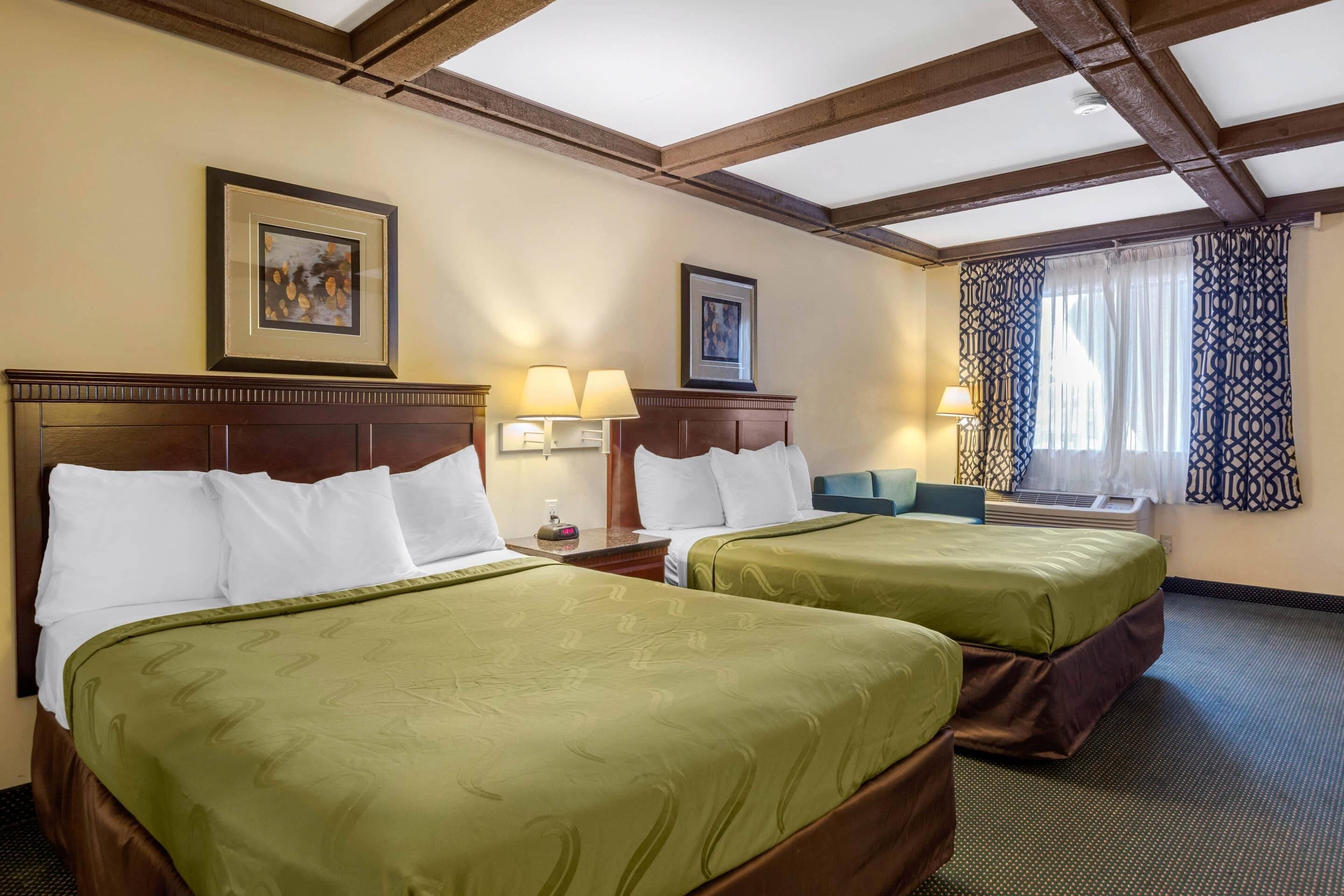 A hotel room with two beds, wooden headboards, framed artwork on the walls, and a window with patterned curtains.