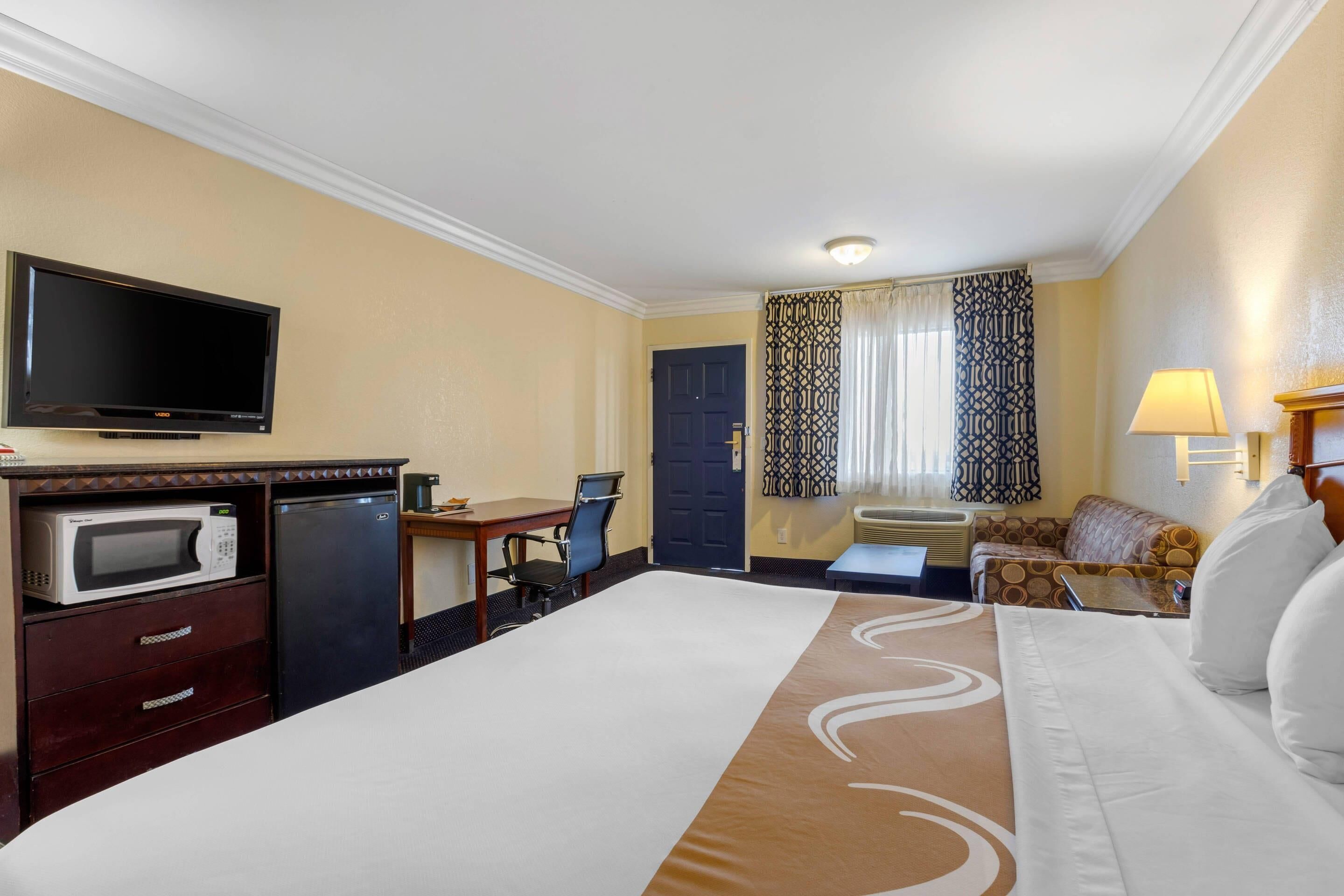 A hotel room with a bed, a desk with a chair, a microwave, a television, and a window with curtains.
