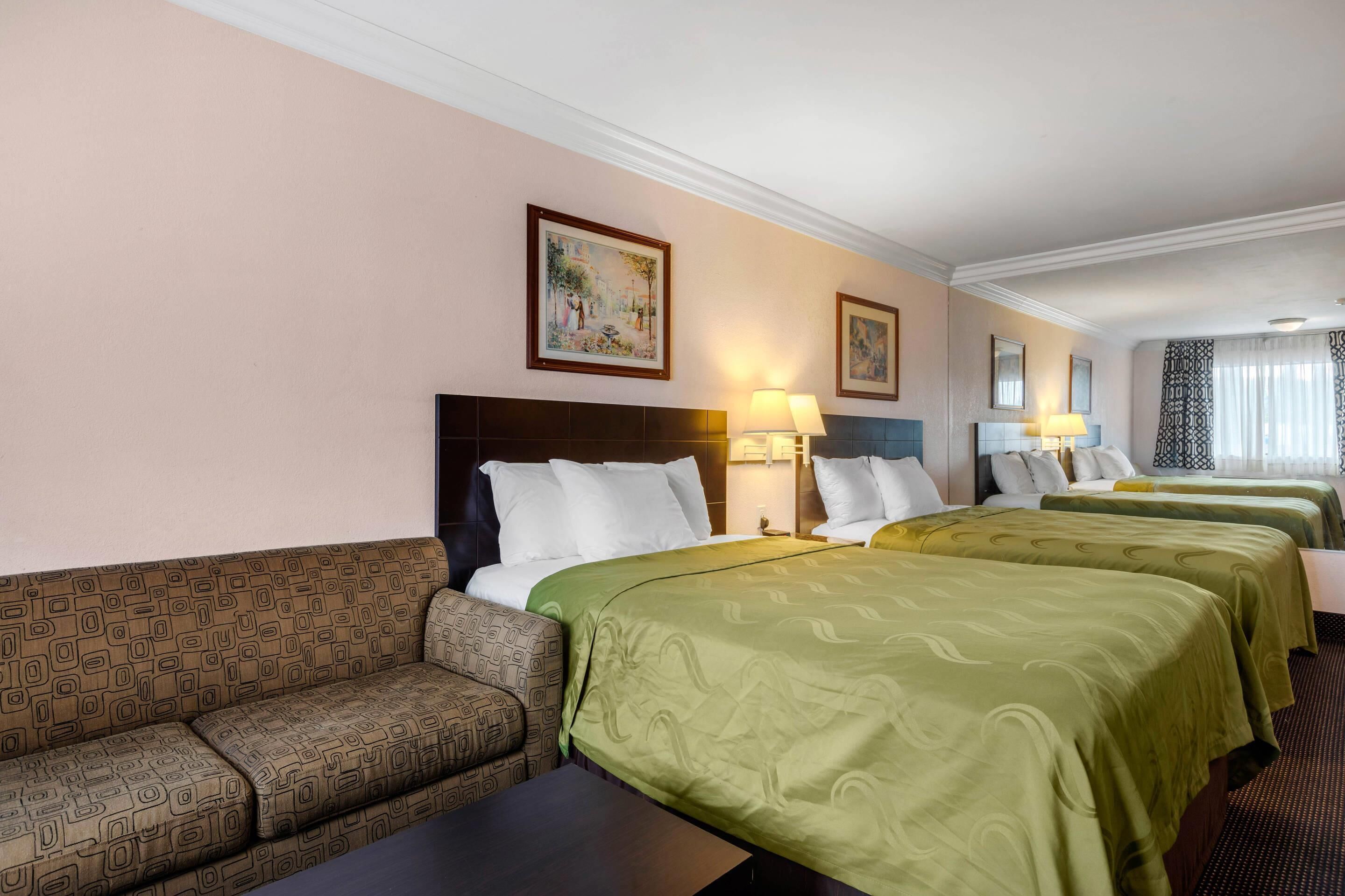Una habitación de hotel con un sofá, dos camas con cubrecamas verdes, una mesita de noche con lámpara y un cuadro en la pared.
