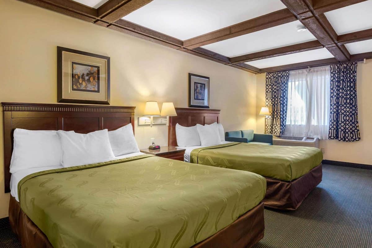 Habitación de hotel con dos camas, cabeceros de madera, lámparas de pared, un cuadro enmarcado y una ventana con cortinas estampadas.