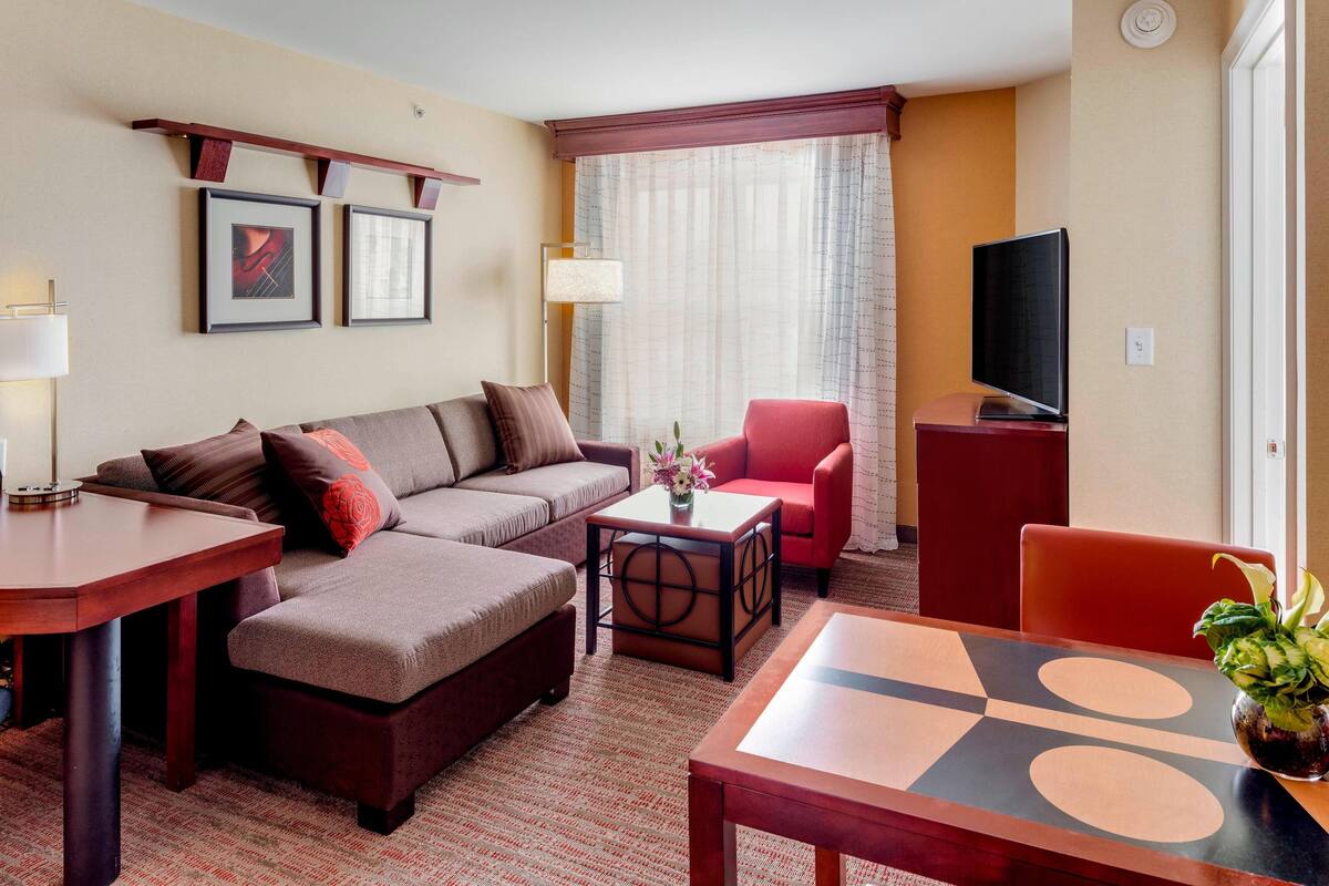 Una habitación de hotel moderna con sofá, reposapiés, una mesita, una silla roja, televisor y una ventana con cortinas.