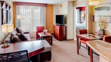 A hotel room with a living area, a dining table, a TV, and a large window with curtains.