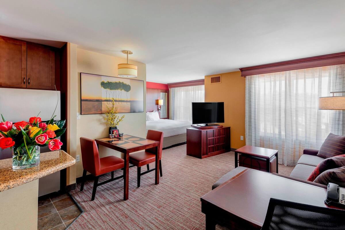 Habitación de hotel con zona de comedor, cama, televisión y ventana con cortinas.