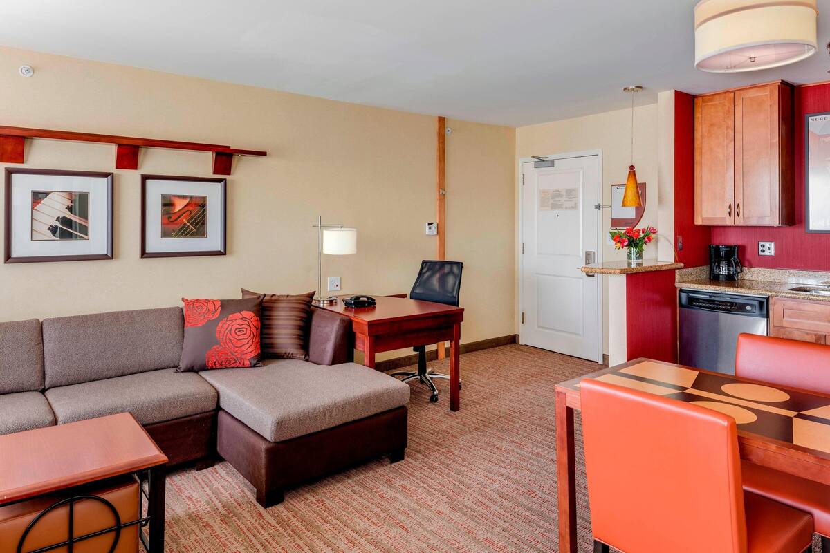 Habitación de hotel compacta con sofá, una pequeña mesa de comedor, zona de cocina con cafetera y refrigerador.