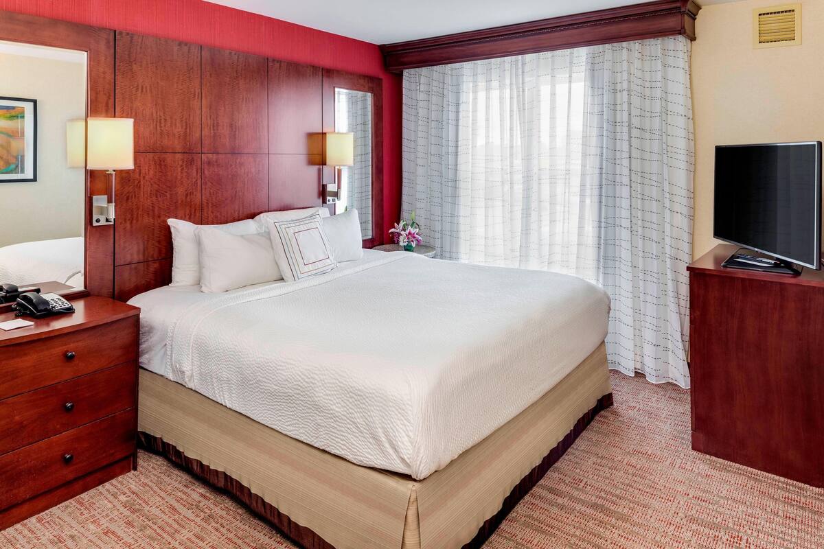 Habitación de hotel con una cama grande, cabecero de madera, una mesita de noche con lámpara, televisor y ventana con cortinas.