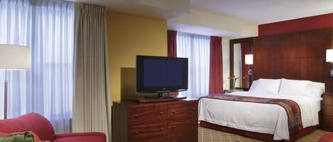 A hotel room with a bed, a TV on a wooden dresser, a red armchair, and a red sofa.