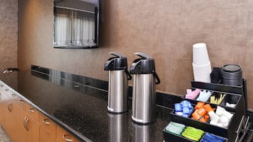 A hotel reception area with a black countertop, two stainless steel coffee makers, a mirror, and a coffee station with disposable cups and utensils.