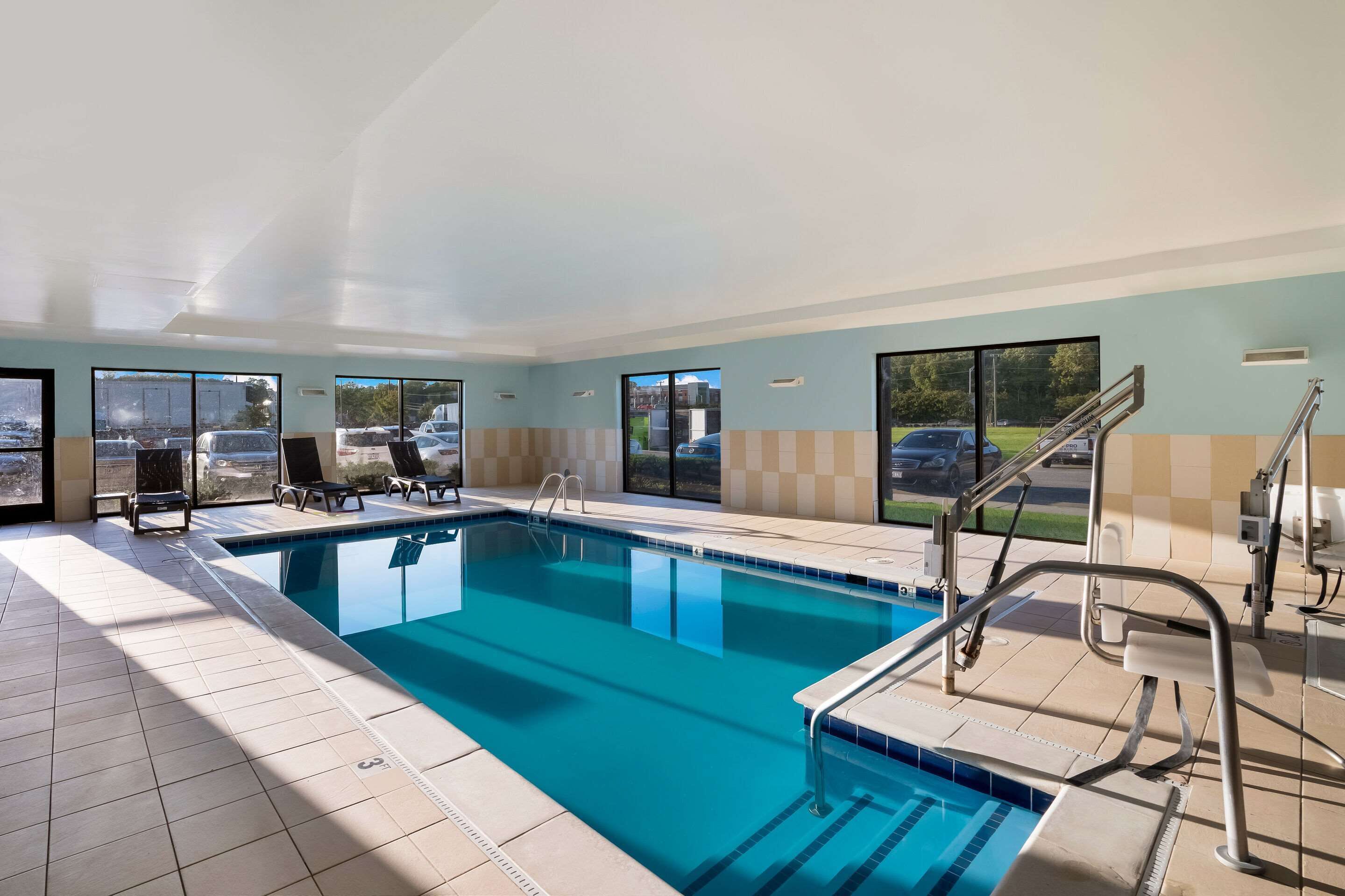 An indoor swimming pool with clear blue water, tiled floor, and a view of the outside through large windows.