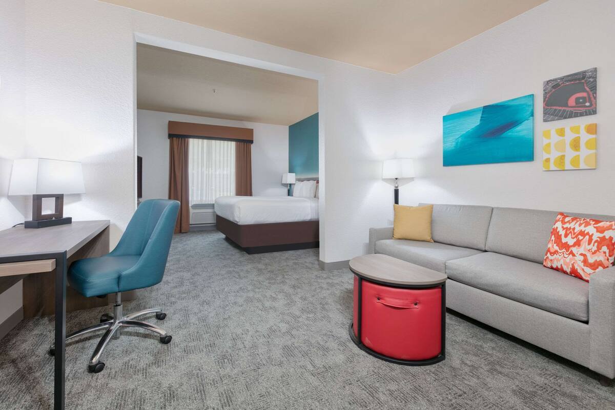 A hotel room with a desk, chair, bed, sofa, and a small red trash bin.