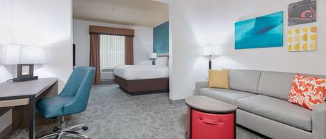 A hotel room with a desk, chair, bed, sofa, and a small red trash bin.