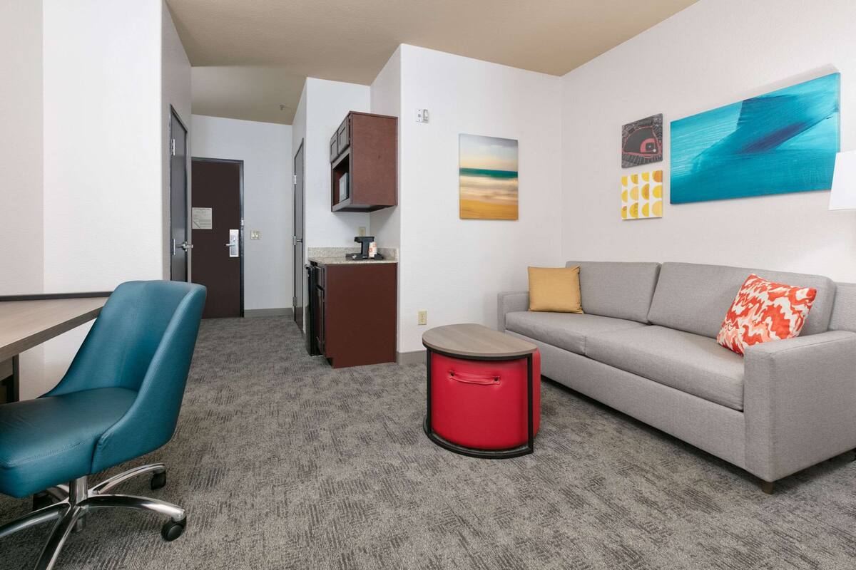 A hotel room with a grey sofa, a blue office chair, a red trash bin, a kitchenette, and a painting on the wall.