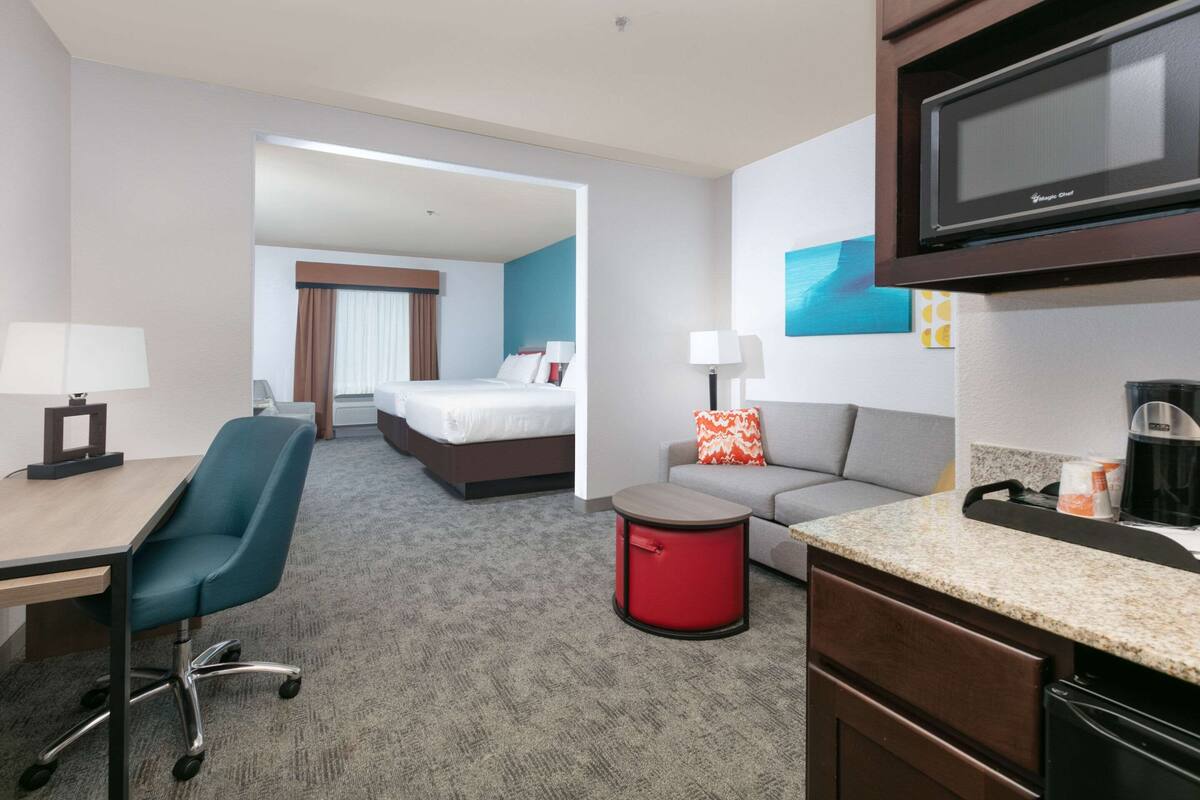 A hotel room with a bed, a desk with a chair, a sofa, a small red stool, a microwave, and a coffee maker.