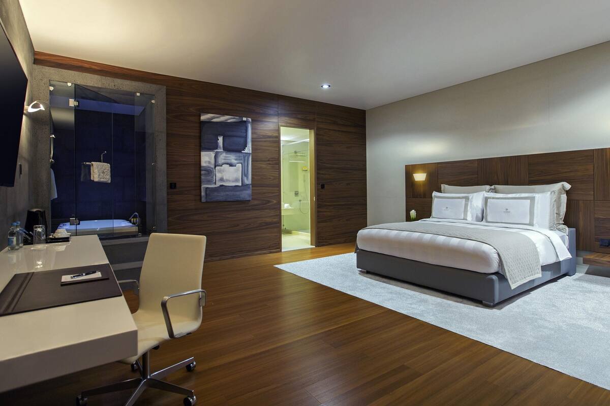 Quarto de hotel moderno com uma cama grande, uma mesa de trabalho com cadeira, um banheiro com espelho e uma parede de destaque em madeira.