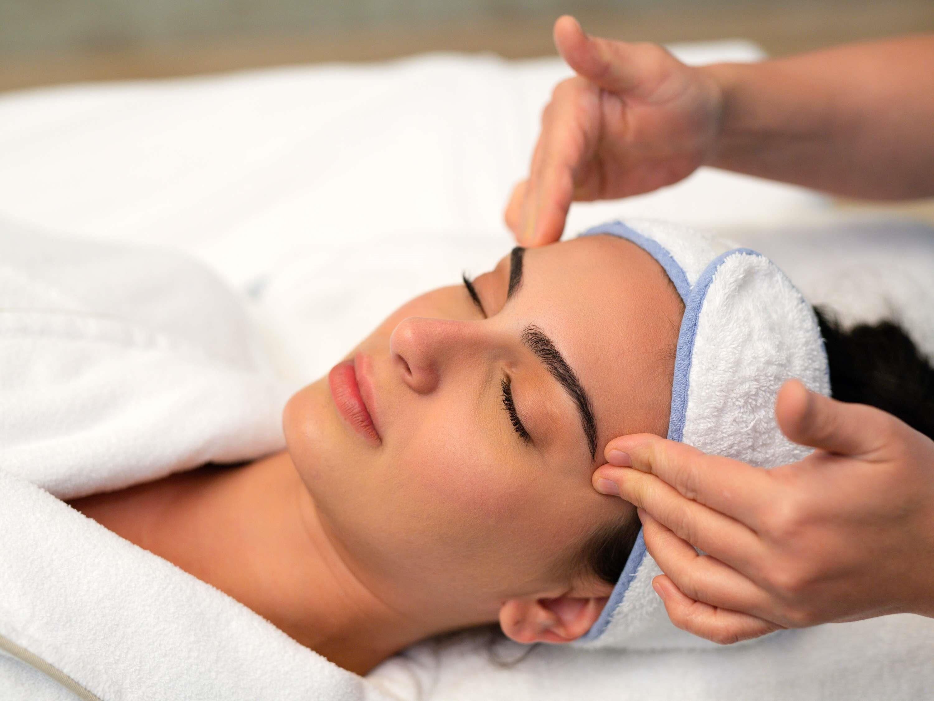 A person lying down with a towel covering their head, and another person applying a treatment to their forehead.