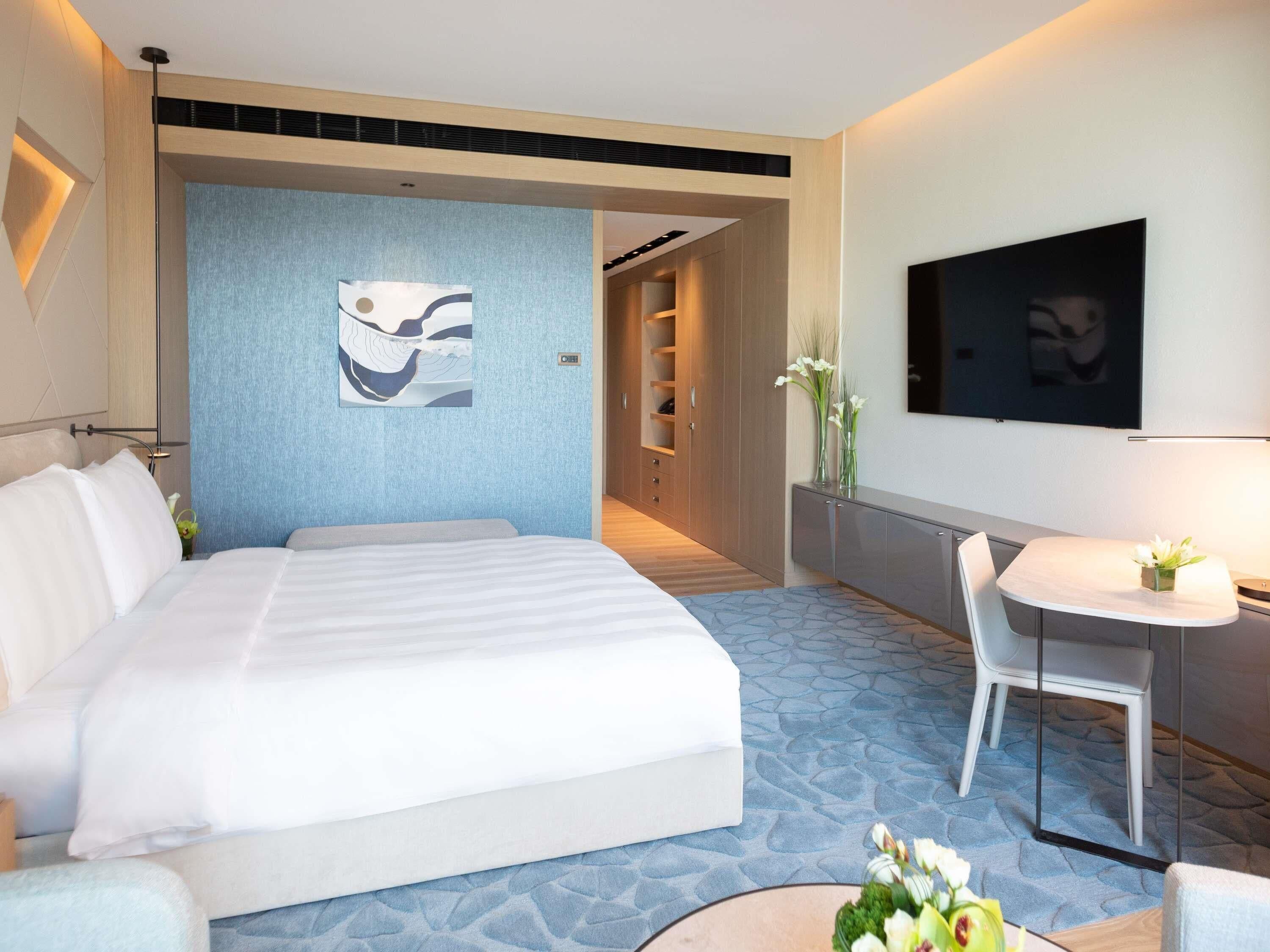 Habitación de hotel con una cama grande, un escritorio con silla, un televisor montado en la pared y un cuadro en la pared.