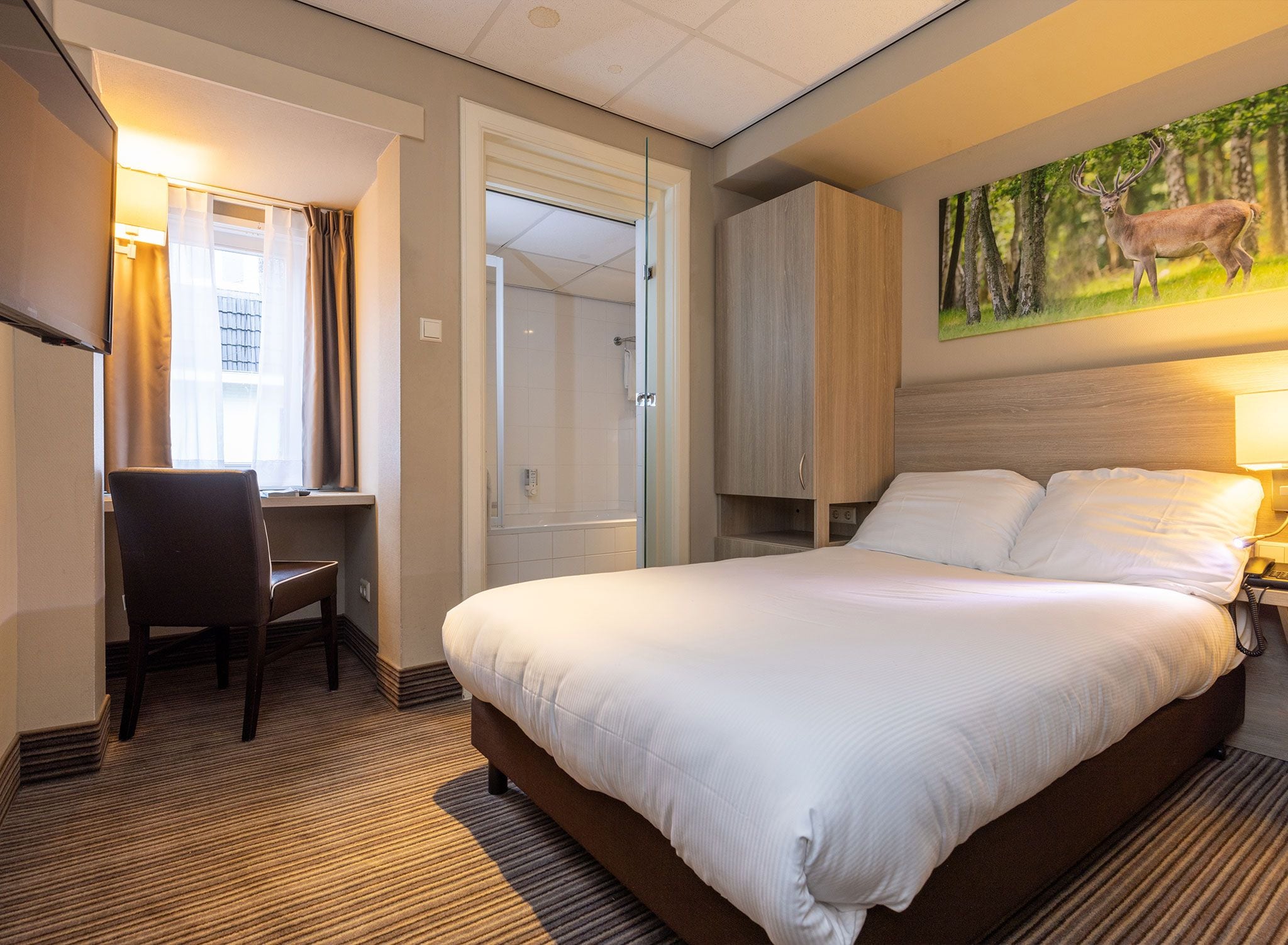 A hotel room with a bed, a desk, a chair, a painting, and a shower area.