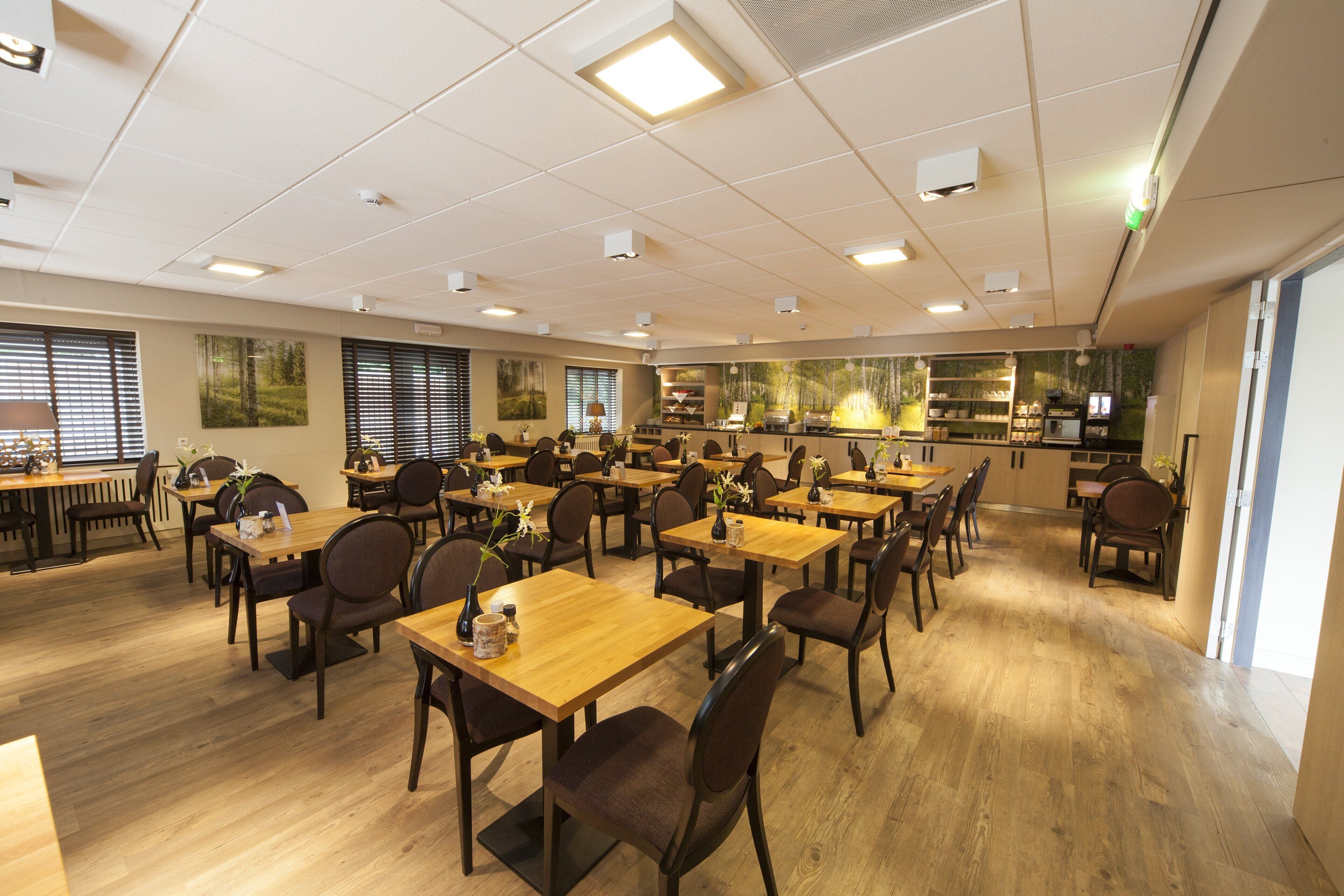 A spacious dining area with multiple tables and chairs, a kitchen in the background, and a view of the outdoors through large windows.