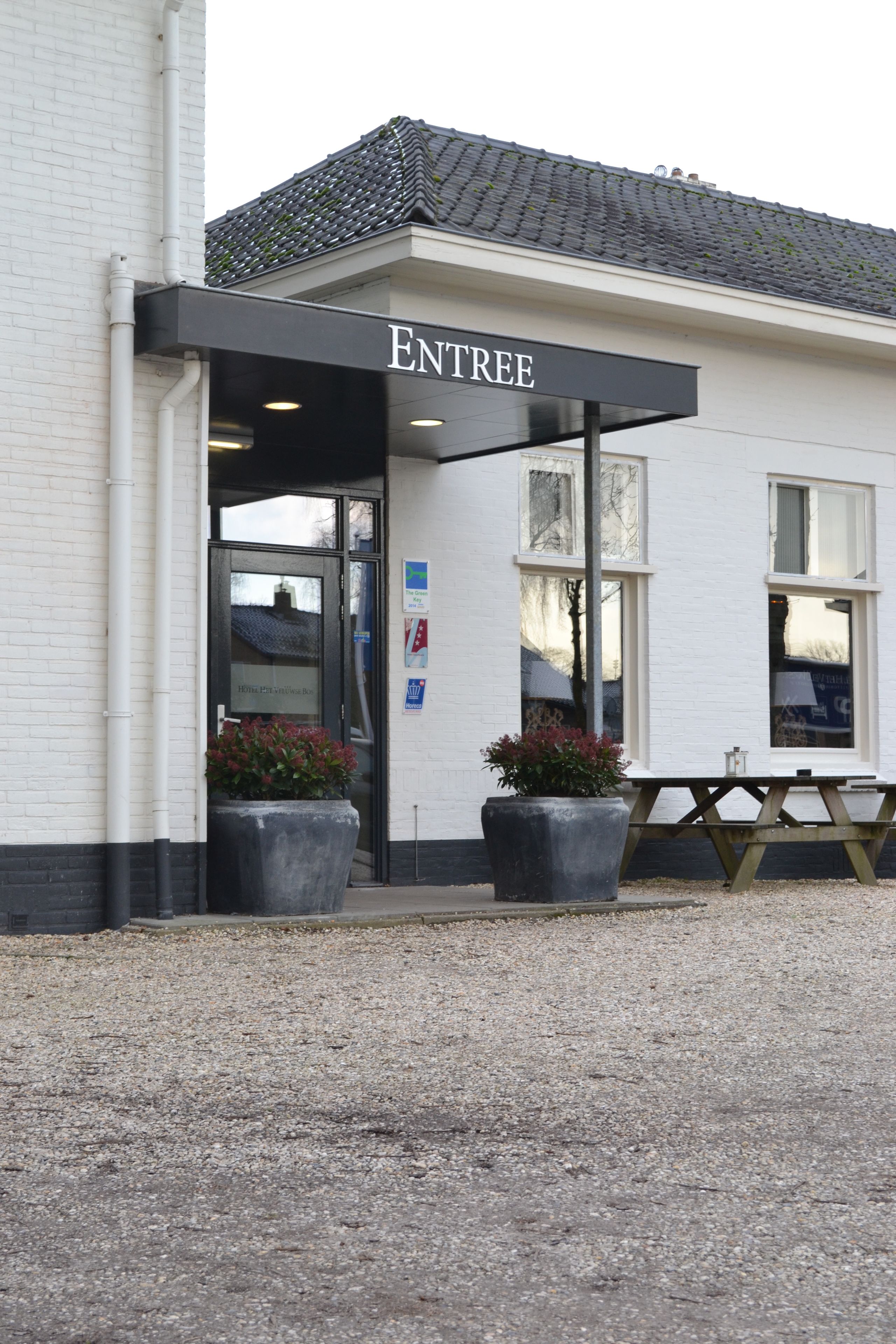 A building with a sign reading "ENTREE" and a covered entrance. There are two large planters with flowers and a picnic table outside.