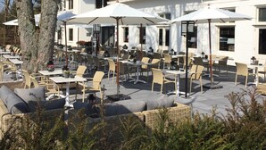 Un café en plein air avec des tables et des chaises, un bâtiment doté d’un toit en pente et un arbre au premier plan.